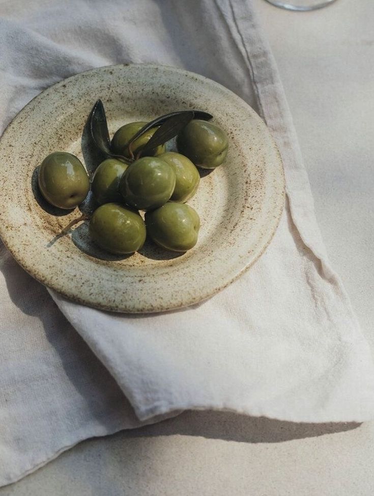 Green olives with dark leaves resting in a small round speckled ceramic dish placed on a light beige cloth