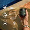 Human hand holding a jar of Ancient Brave True Magnesium supplement next to a glass of water on a golden colored table, with information about product benefits