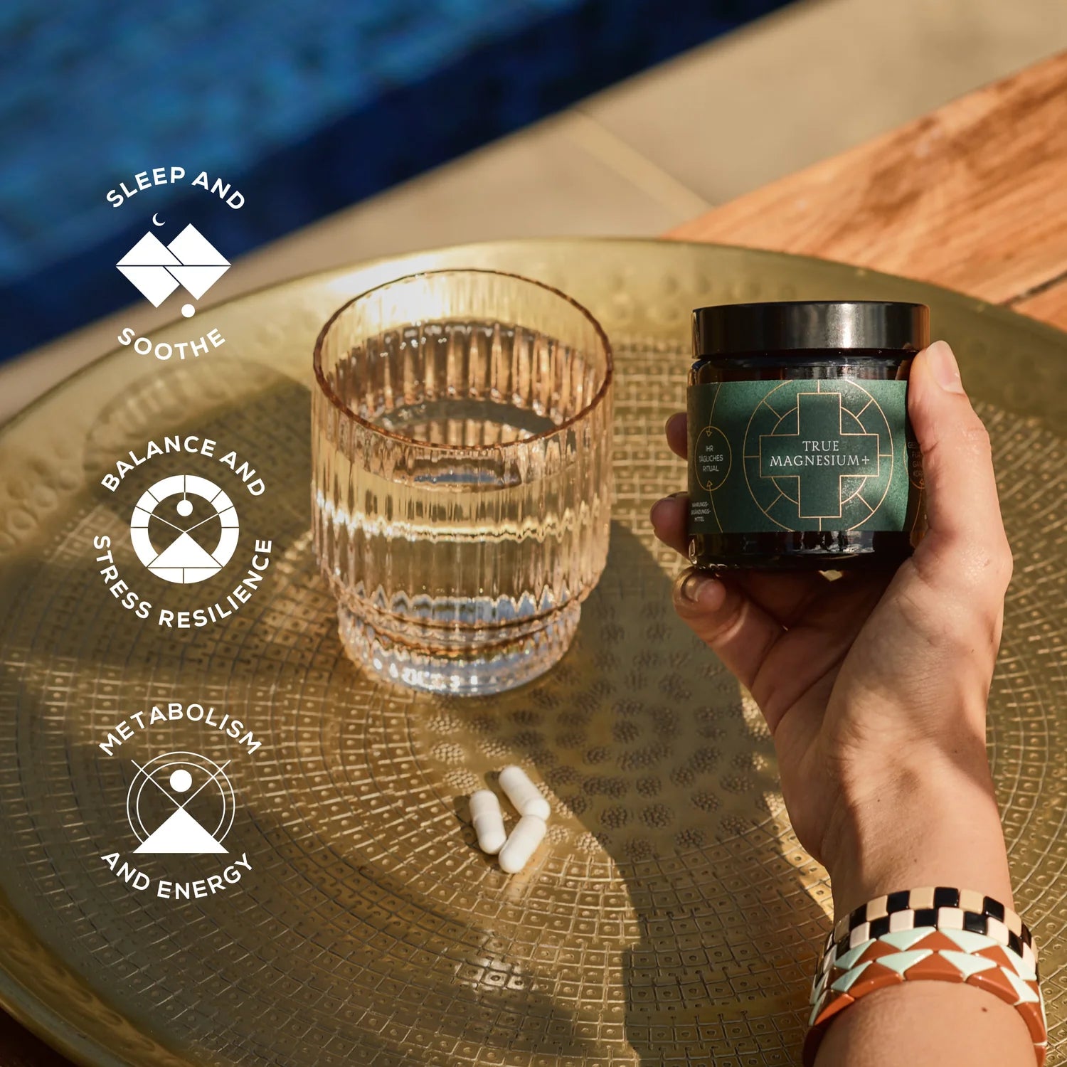 Human hand holding a jar of Ancient Brave True Magnesium supplement next to a glass of water on a golden colored table, with information about product benefits